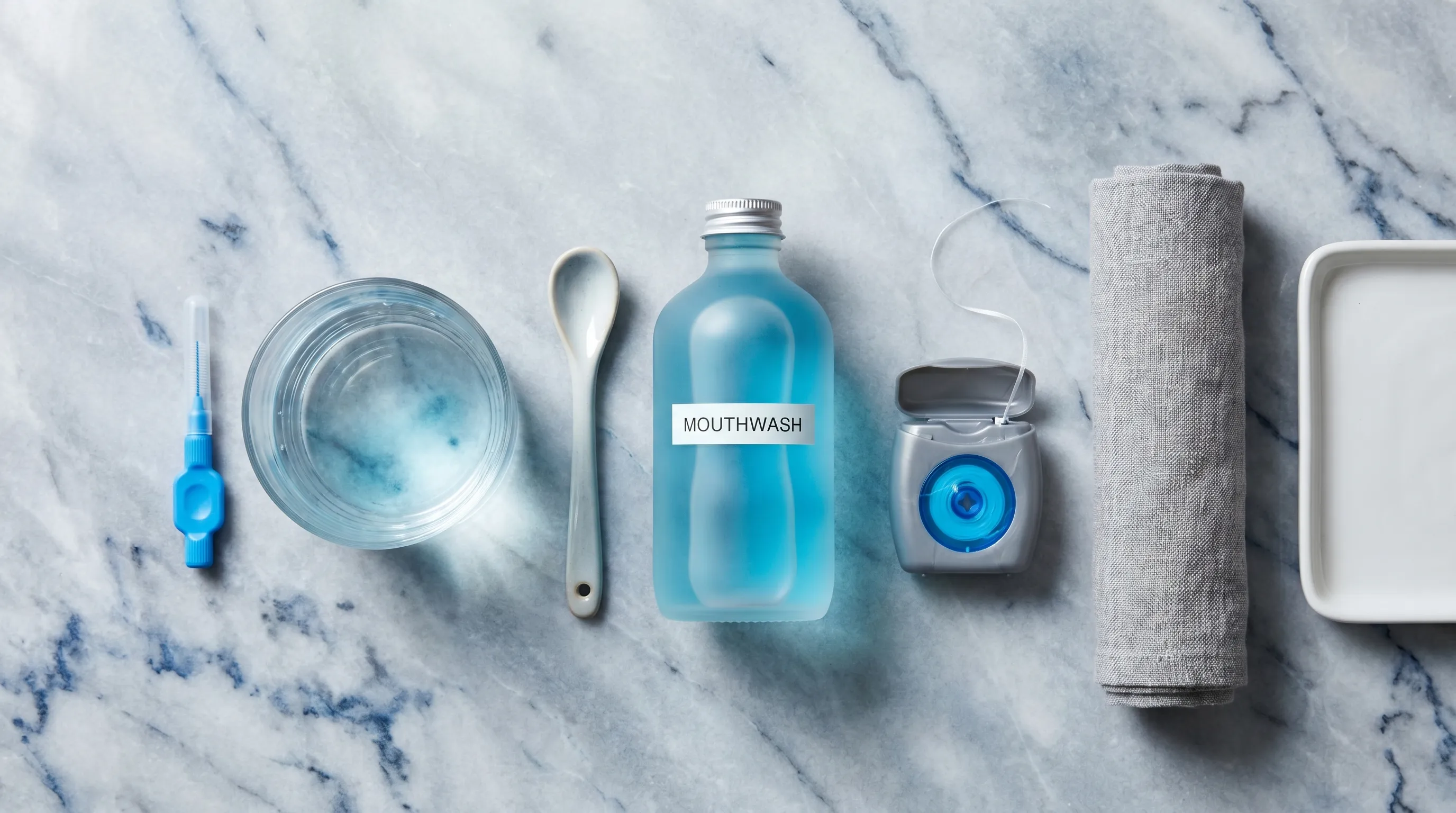 Overhead flatlay of essential oral hygiene tools including an interdental brush, saltwater rinse glass, antibacterial mouthwash, and dental floss on a clean marble countertop