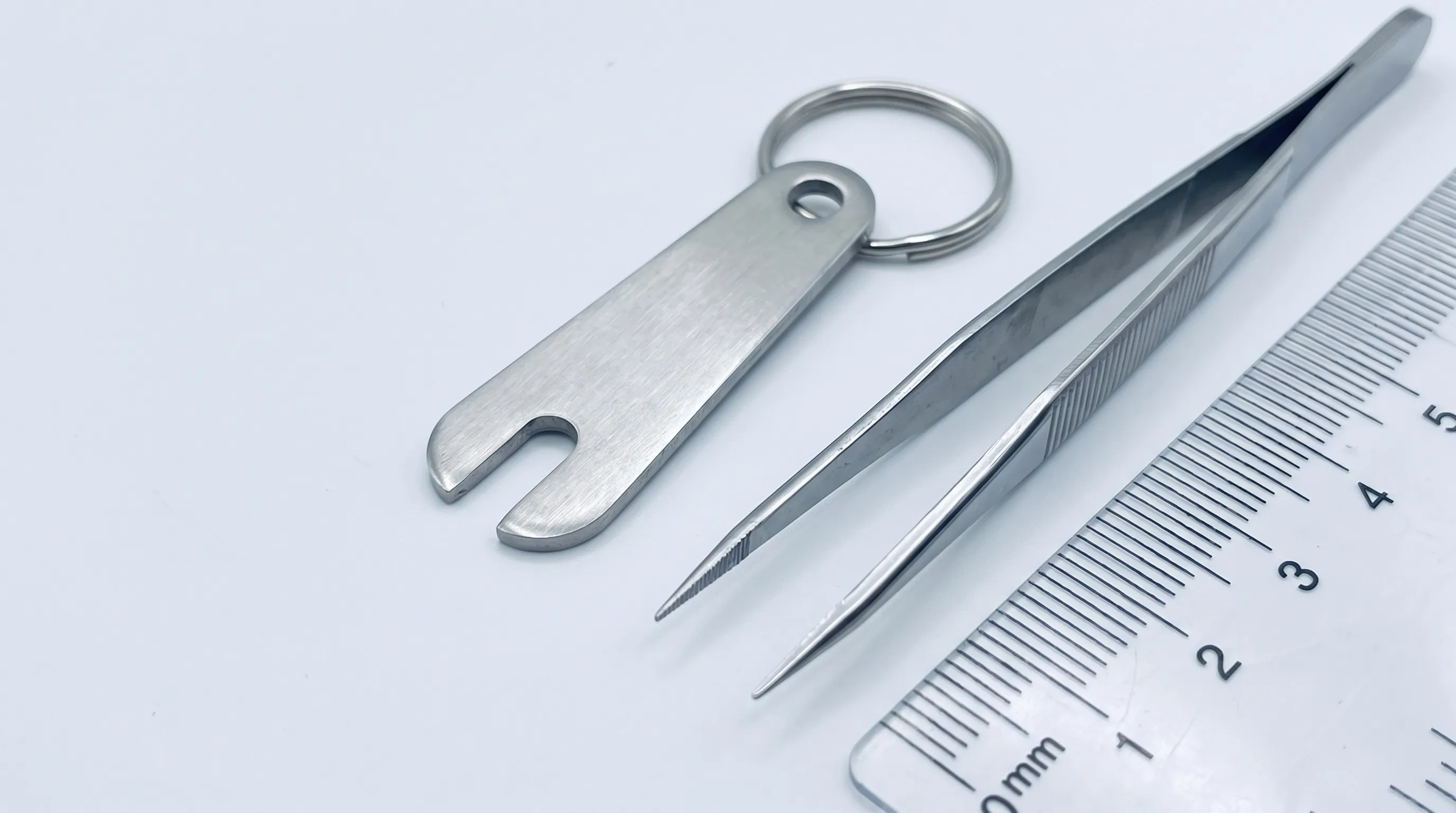 A close-up medical photography shot of a stainless steel tick removal keychain next to a standard pair of pointed tweezers on a clean clinical white background, highlighting the tool's ergonomic notch design and compact size