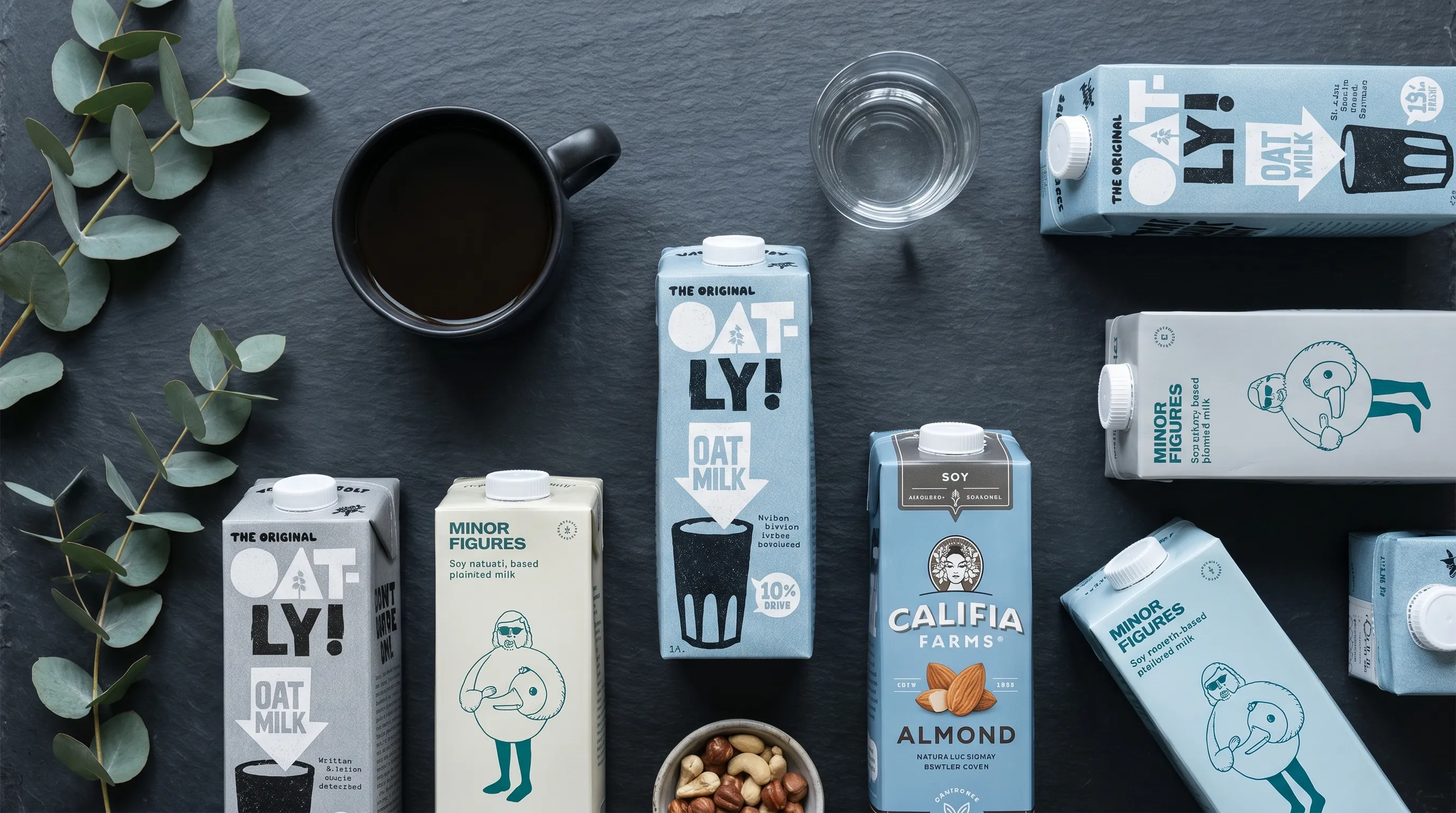 A minimalist flatlay showing black coffee, a glass of water, and various plant-based milk cartons arranged on a slate surface, emphasizing healthy lifestyle choices with cool blue and gray tones.