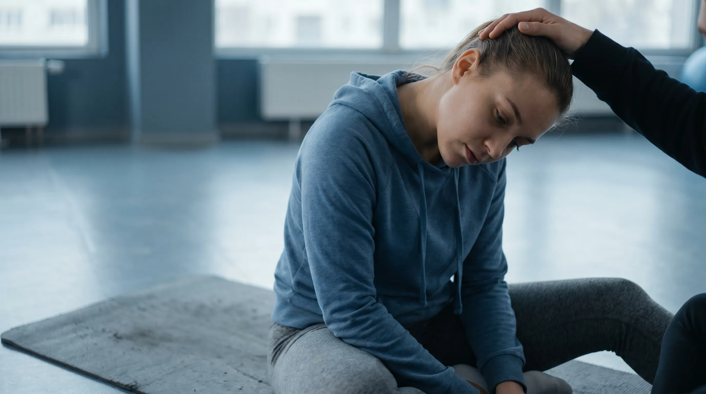 A close-up photograph of a person in athletic wear gently stretching their upper trapezius muscle on a yoga mat, gray and blue tones, soft focus background, 4:3 ratio